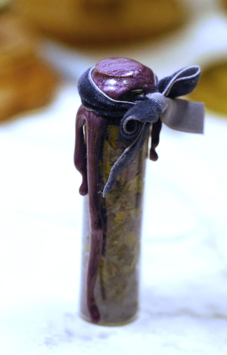 May include: A small glass vial filled with dried herbs, sealed with a dark purple stopper. The stopper is adorned with a dark gray velvet ribbon tied in a bow, and a matching tag. The vial is decorated with dripping purple wax.