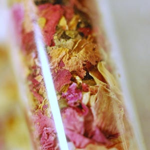 May include: A clear glass vial filled with a mix of dried flowers and herbs. The contents include pink rose petals, beige and brown dried leaves, and small white granules. The vial is angled, with a bright white reflection.