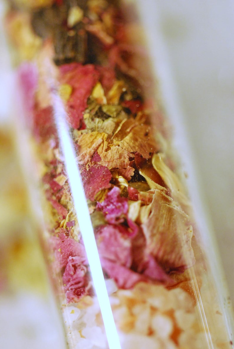 May include: A clear glass vial filled with a mix of dried flowers and herbs. The contents include pink rose petals, beige and brown dried leaves, and small white granules. The vial is angled, with a bright white reflection.