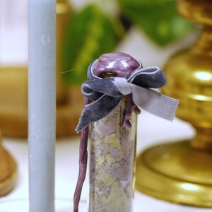 May include: A small glass vial filled with dried botanicals, sealed with a purple wax and tied with a dark blue velvet ribbon. A light blue candle stands to the left. The background includes a gold candlestick and greenery.