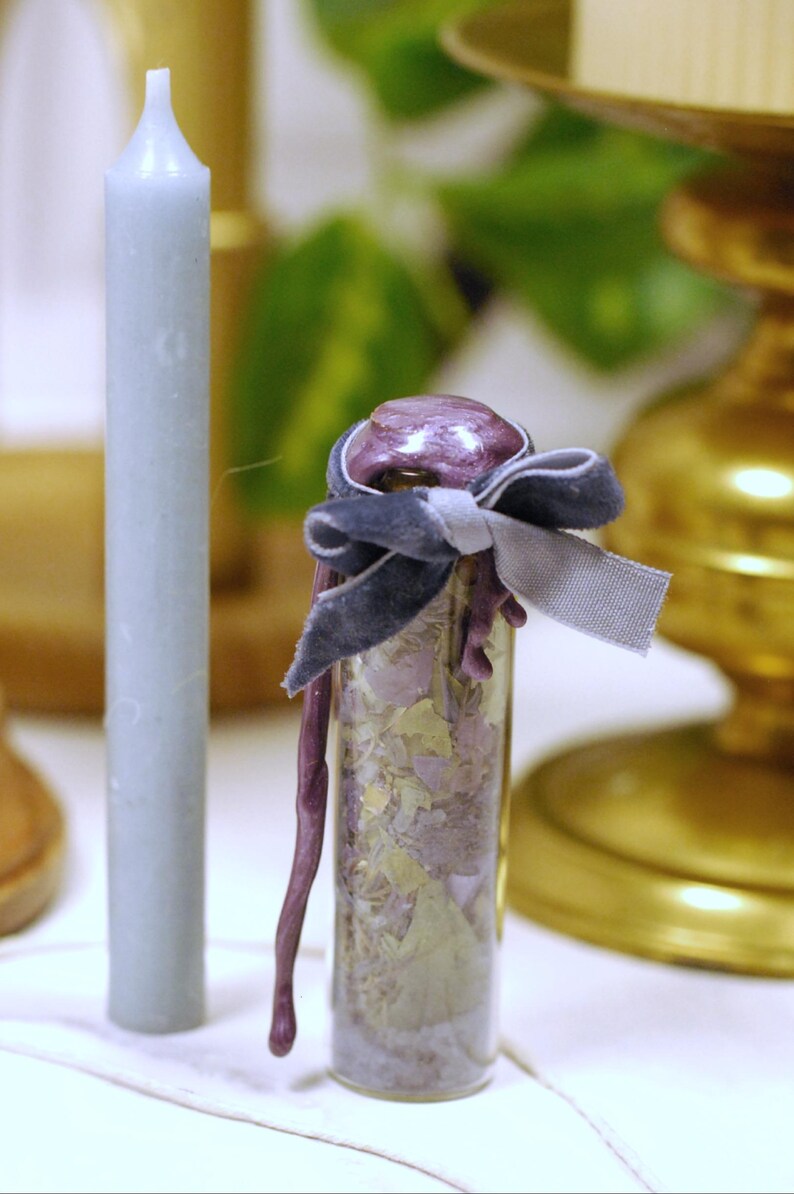 May include: A small glass vial filled with dried botanicals, sealed with a purple wax and tied with a dark blue velvet ribbon. A light blue candle stands to the left. The background includes a gold candlestick and greenery.