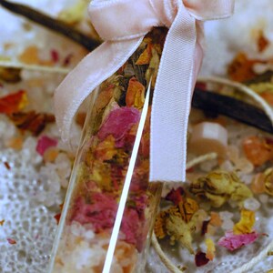 May include: A small glass vial filled with layers of pink and orange dried flower petals, white crystals, and other botanicals. The vial is tied with a light pink velvet ribbon. A vanilla bean and other decorative elements are in the background.