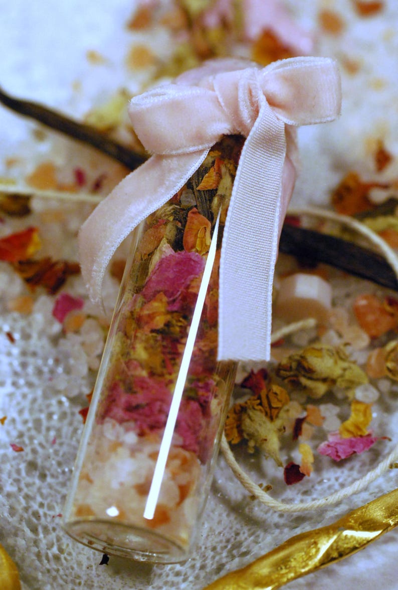 May include: A small glass vial filled with layers of pink and orange dried flower petals, white crystals, and other botanicals. The vial is tied with a light pink velvet ribbon. A vanilla bean and other decorative elements are in the background.