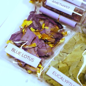 May include: Close-up of several small clear glass bottles and clear plastic bags filled with dried botanicals. The bags are labeled with the names of the contents: Blue Lotus, Eucalyptus, and Amethyst. The Blue Lotus has purple and yellow petals.