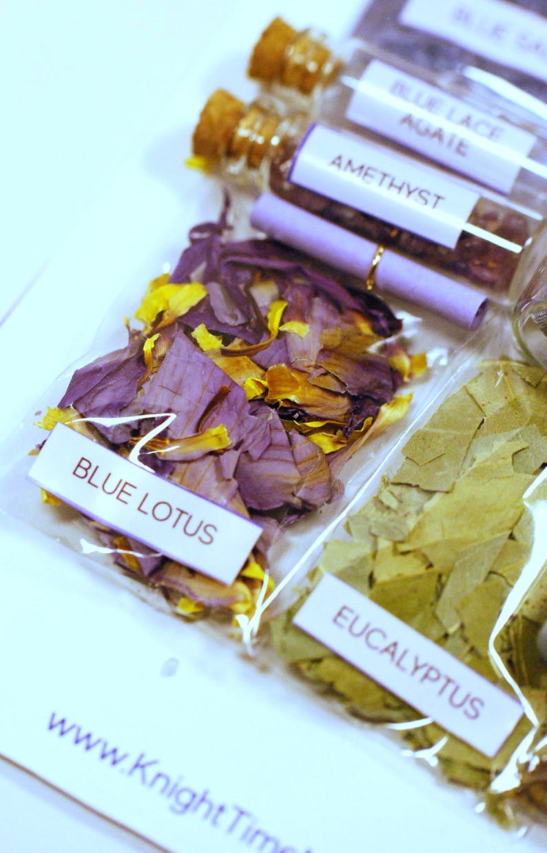 May include: Close-up of several small clear glass bottles and clear plastic bags filled with dried botanicals. The bags are labeled with the names of the contents: Blue Lotus, Eucalyptus, and Amethyst. The Blue Lotus has purple and yellow petals.