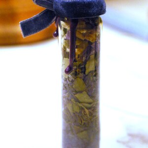May include: A small glass bottle filled with dried herbs and a dark purple wax seal. The bottle is topped with a dark blue velvet ribbon tied in a bow. The wax seal drips down the side of the bottle.