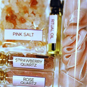 May include: An assortment of self-care products, including pink salt, strawberry quartz, rose quartz, and ritual oil. The items are packaged in clear containers and bags, with labels identifying each product. A glass jar contains pink ribbon.