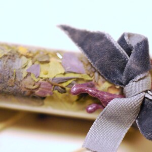 May include: A clear glass vial filled with dried herbs and flowers, tied with a gray velvet ribbon. The vial's contents include green, yellow, and purple plant matter. A small, purple, decorative element is attached to the vial.
