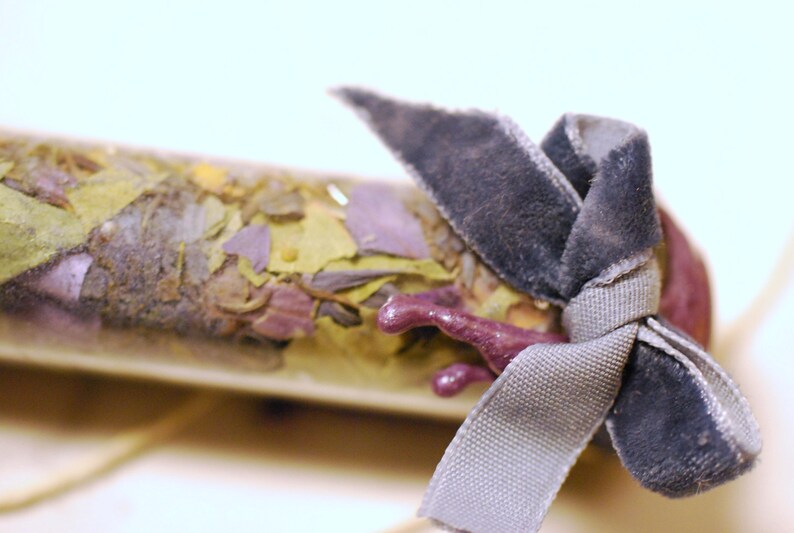 May include: A clear glass vial filled with dried herbs and flowers, tied with a gray velvet ribbon. The vial's contents include green, yellow, and purple plant matter. A small, purple, decorative element is attached to the vial.