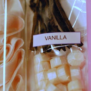 May include: A clear glass bottle with pink ribbon and a package of vanilla beans and wax melts. The label on the package reads "VANILLA". The vanilla beans are dark brown and the wax melts are white.