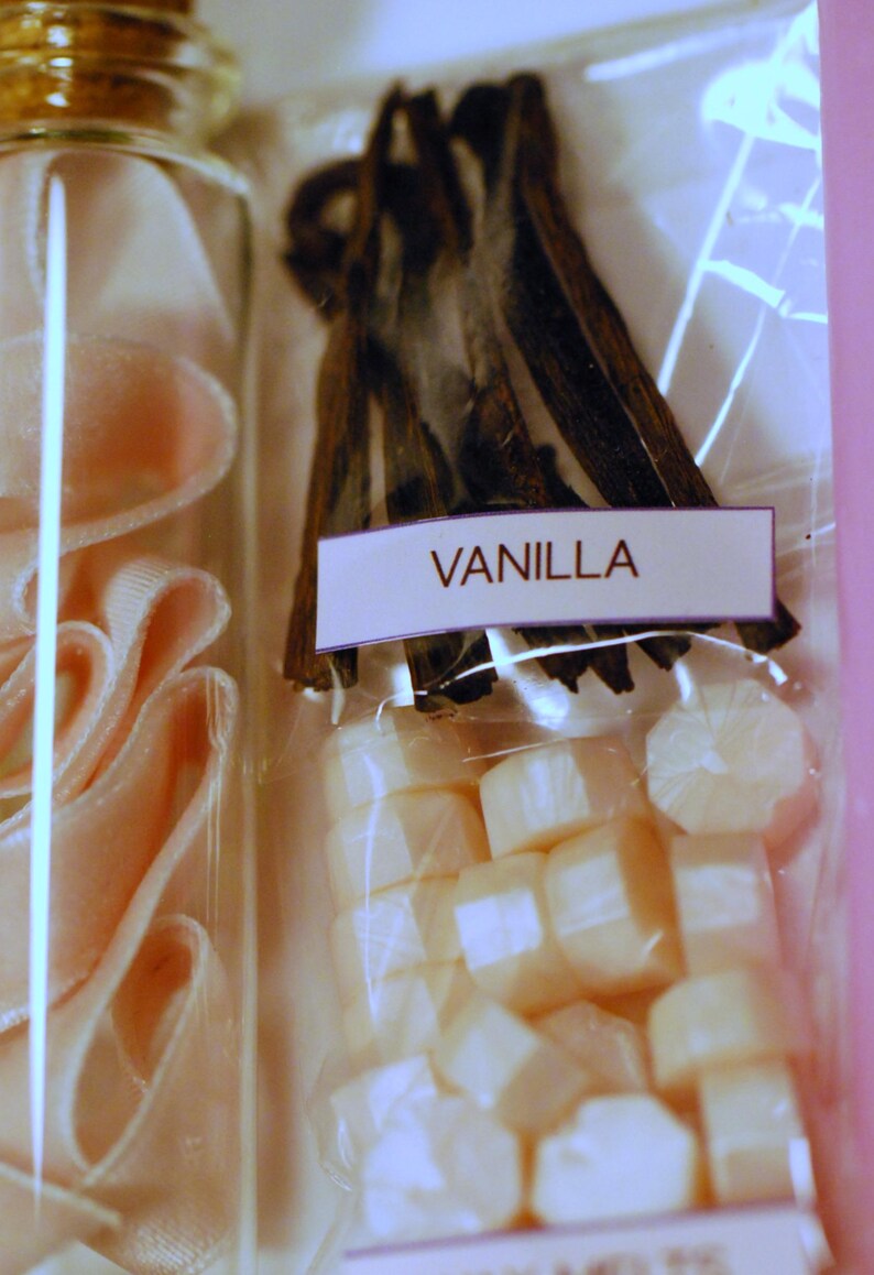 May include: A clear glass bottle with pink ribbon and a package of vanilla beans and wax melts. The label on the package reads "VANILLA". The vanilla beans are dark brown and the wax melts are white.