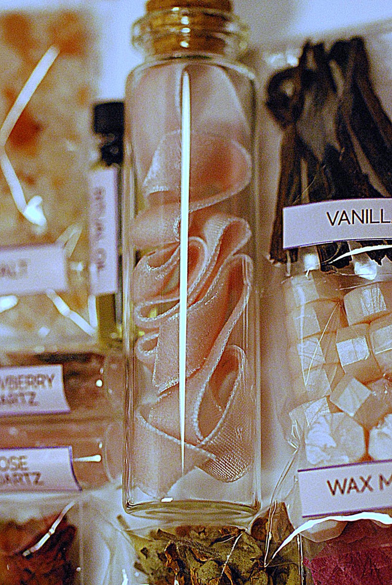 May include: A clear glass bottle filled with pink ribbon, next to a package of vanilla beans and wax melts. The bottle has a cork stopper. Other small bottles and packages are visible in the background.