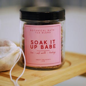Puede incluir: Un frasco de vidrio transparente con tapa negra, lleno de una mezcla de té de baño botánico. Una etiqueta rosa dice "SOAK IT UP BABE" y "Botanical Bath Tea Blend". Una pequeña bolsa de muselina está al lado del frasco en una bandeja de madera.