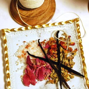May include: A decorative arrangement featuring a white plate with a gold-colored twisted border. The plate holds pink rose petals, vanilla beans, and salt crystals. A wooden spool of twine sits above.