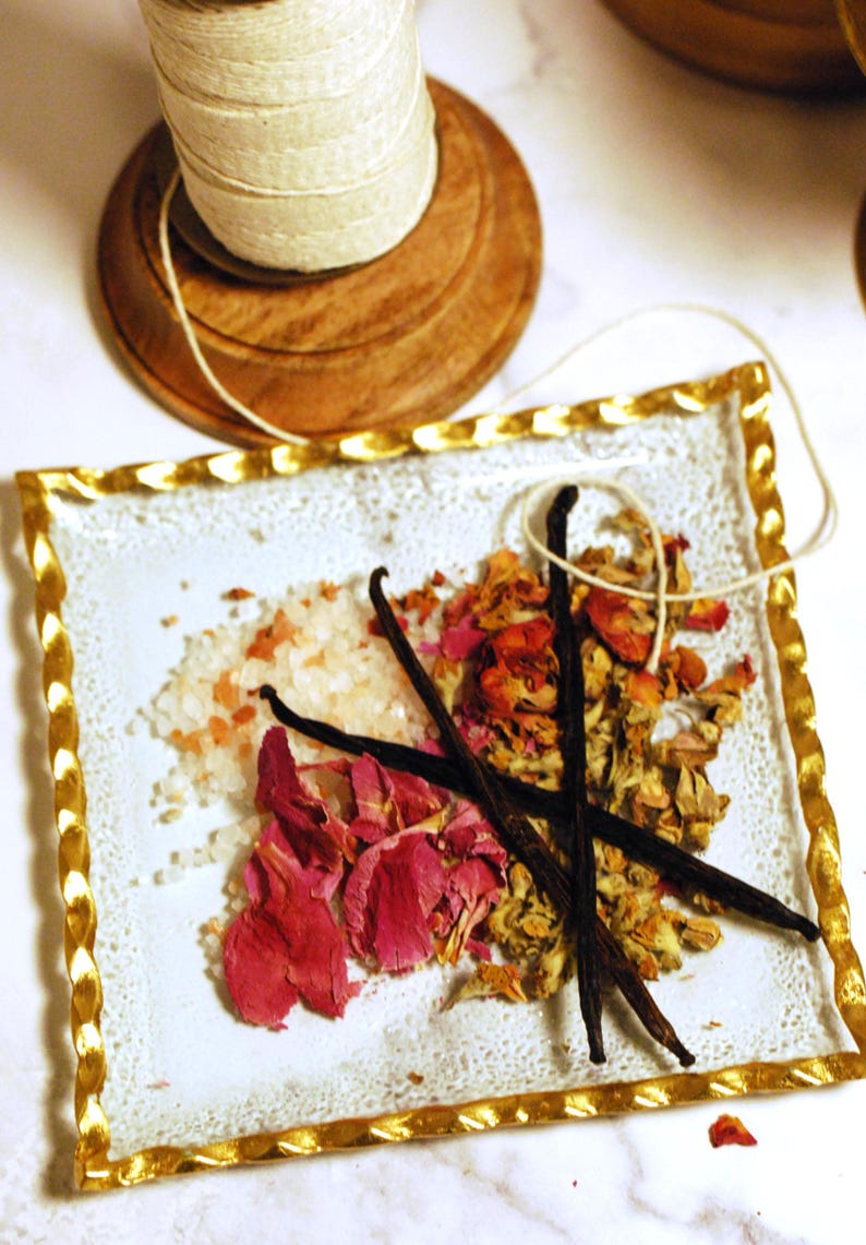 May include: A decorative arrangement featuring a white plate with a gold-colored twisted border. The plate holds pink rose petals, vanilla beans, and salt crystals. A wooden spool of twine sits above.