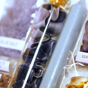 May include: A clear glass bottle filled with navy blue velvet ribbon and a cork stopper. A light blue candle is next to the bottle. The image also includes a package with seashells.