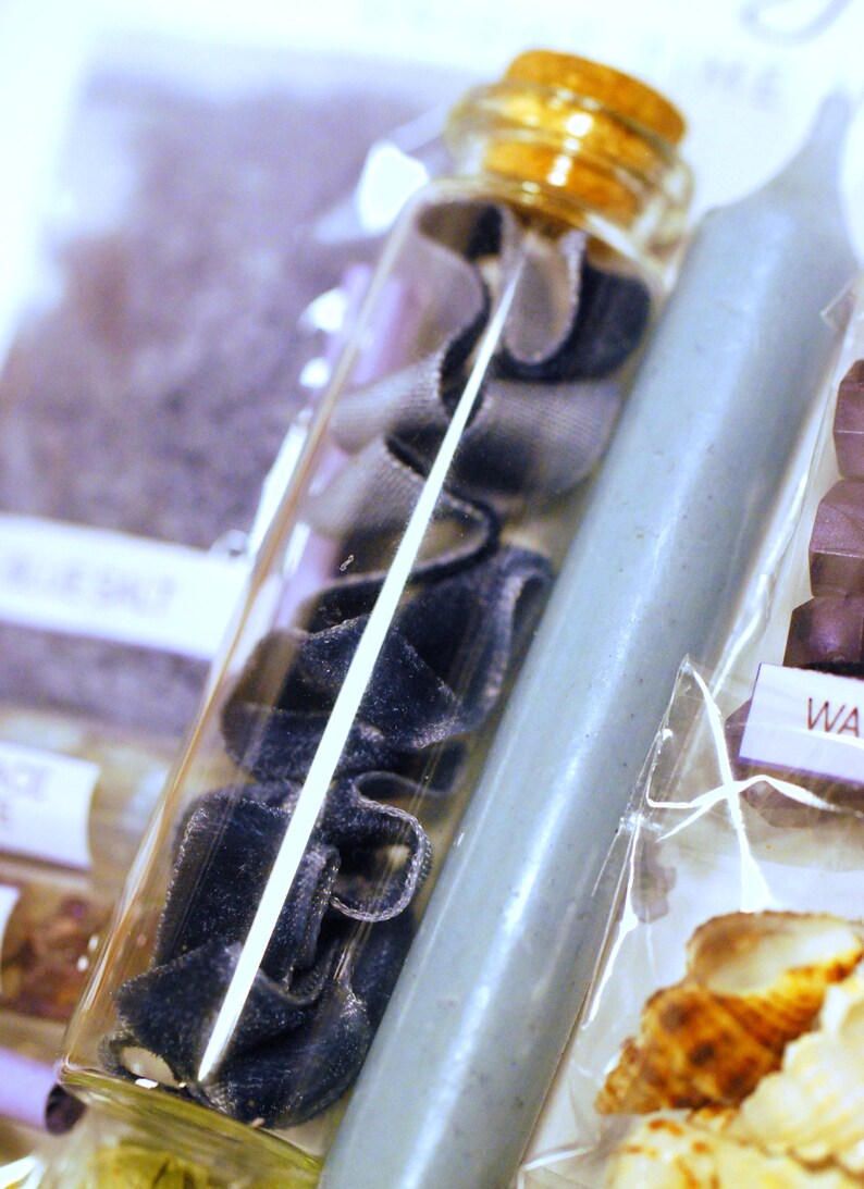 May include: A clear glass bottle filled with navy blue velvet ribbon and a cork stopper. A light blue candle is next to the bottle. The image also includes a package with seashells.