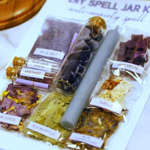 May include: A DIY spell jar kit for anxiety, featuring various ingredients in clear packaging. Includes blue salt, amethyst, blue lace agate, wax melts, shells, and a gray candle. The kit also contains dried flowers and eucalyptus leaves. The text "DIY SPELL JAR KIT" is visible.