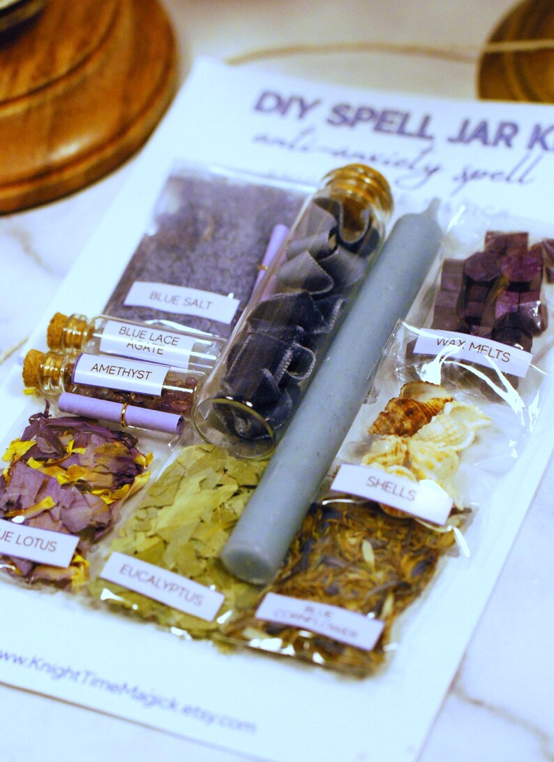 May include: A DIY spell jar kit for anxiety, featuring various ingredients in clear packaging. Includes blue salt, amethyst, blue lace agate, wax melts, shells, and a gray candle. The kit also contains dried flowers and eucalyptus leaves. The text "DIY SPELL JAR KIT" is visible.