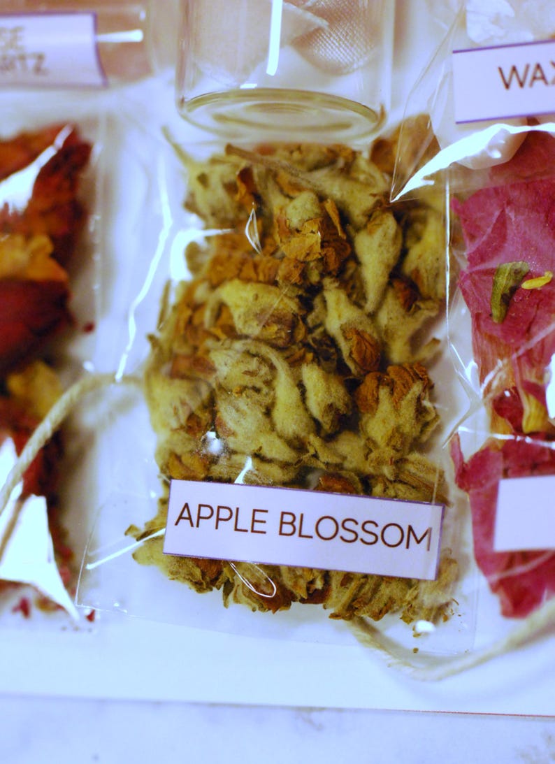 May include: Close-up of dried apple blossoms in a clear plastic bag. The label reads "APPLE BLOSSOM". The blossoms are a mix of light brown and yellow. Other bags with dried flowers are visible.