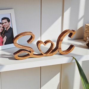 Puede incluir: Letrero decorativo de madera con las letras "S" y "C" unidas por un corazón calado. El letrero es de madera marrón claro y está sobre una estantería blanca. En el fondo se ve una foto enmarcada.