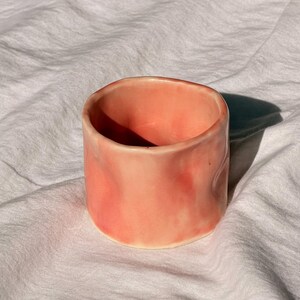 May include: A small, cylindrical ceramic cup in a soft, peachy-pink hue. The cup has a smooth, glossy finish with subtle variations in colour, creating a textured appearance. The open top and rounded edges give it a simple, modern design.