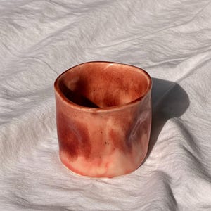 May include: A handmade ceramic cup with a reddish-pink glaze. The cup has a cylindrical shape with a slightly irregular rim and a smooth, textured surface. The color transitions from a lighter pink at the base to a deeper red at the top, with subtle variations in tone.