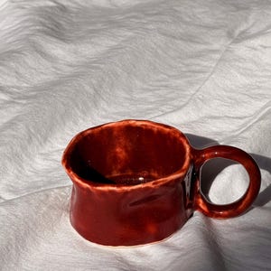 May include: A small, handmade ceramic mug in a deep burgundy colour. The mug has a rounded body, a small handle, and a textured surface. The interior is dark, and the mug sits on a white, textured surface.