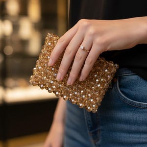 Könnte beinhalten: Eine goldfarbene, perlenbesetzte Clutch mit rechteckiger Form. Die Clutch wird von einer Person gehalten, die einen goldenen Ring mit einem quadratischen Diamanten trägt. Die Person trägt ein schwarzes Oberteil und blaue Jeans.