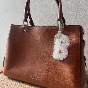 May include: A brown leather handbag with a beaded charm in the shape of the letter "B" hanging from the handle. The handbag has two handles and a silver-tone clasp. The charm is made of iridescent beads.