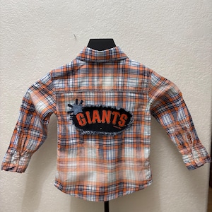 May include: Orange, white, and blue plaid button-down shirt with long sleeves. A black patch on the back features the word "GIANTS" in orange lettering. The shirt is displayed on a black hanger.