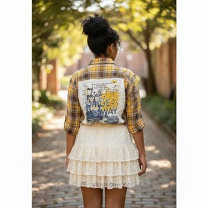 May include: A yellow and brown plaid long-sleeve button-down shirt featuring a white rectangular patch on the back with the text 'my perfect HIDE AWAY' in black and yellow ink, paired with a cream-colored tiered lace mini skirt.