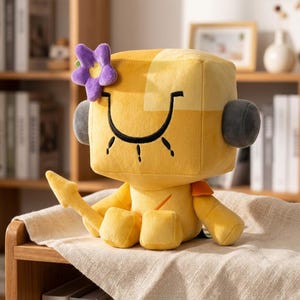Puede incluir: Un peluche amarillo con forma de cubo que presenta una gran sonrisa bordada en negro, accesorios laterales grises en forma de orejas circulares y una flor morada en la parte superior izquierda. El personaje tiene una cola amarilla puntiaguda.