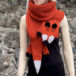 May include: A knitted orange scarf with a fox design. The scarf features a fox face with black and white accents, and the tail has a white tip. The scarf is displayed on a mannequin against a stone wall background.