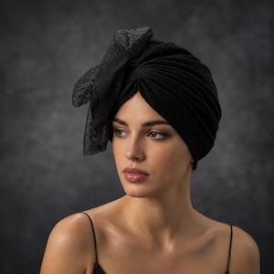Może przedstawiać: Czarny turban z plisowanym wzorem i dużą, ozdobną kokardą z boku, wykonaną z przezroczystej tkaniny w kropki. Nakrycie głowy wykonane jest z teksturowanego, ciemnego materiału, kt&oacute;ry zakrywa czubek i boki głowy, tworząc ustrukturyzowaną formę.