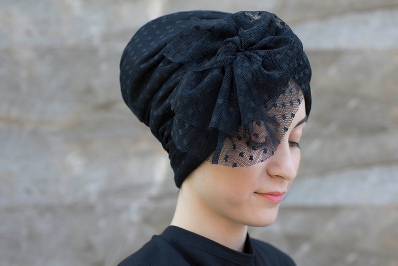 Może przedstawiać: Czarny turban wykonany z teksturowanego materiału z subtelnym wzorem w gwiazdki. Projekt zawiera dużą, warstwową kokardę z przezroczystej, kropkowanej siateczki umieszczoną z boku, z częścią siateczki opadającą na czoło.