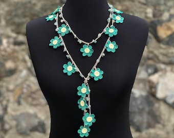 Può includere: Una collana a due fili all'uncinetto con fiori verdi e bianchi. La collana è realizzata con un cordoncino beige e ha un lungo ciondolo con più fiori verdi e bianchi.