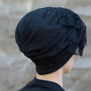 Może przedstawiać: Czarny turban wykonany z przezroczystej siateczki w kropki. Projekt charakteryzuje się marszczonymi fałdami oraz warstwowym, falbankowym akcentem z boku, co nadaje mu teksturę. Warstwa podstawowa wykonana jest z gładkiego, czarnego materiału.