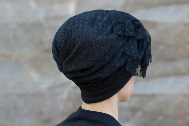 Może przedstawiać: Czarny turban wykonany z przezroczystej siateczki w kropki. Projekt charakteryzuje się marszczonymi fałdami oraz warstwowym, falbankowym akcentem z boku, co nadaje mu teksturę. Warstwa podstawowa wykonana jest z gładkiego, czarnego materiału.