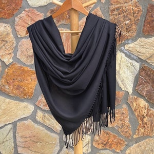 Puede incluir: Un chal de pashmina negro con flecos. El chal está drapeado sobre una percha de madera y se muestra contra una pared de piedra.