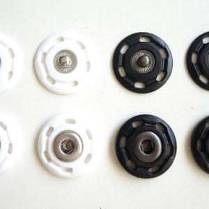 May include: Six snap fasteners, three white and three black. Each fastener has a metal center and a plastic outer ring with a decorative pattern.