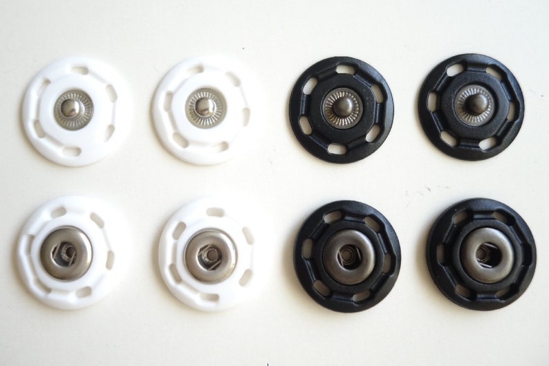 May include: Six snap fasteners, three white and three black. Each fastener has a metal center and a plastic outer ring with a decorative pattern.