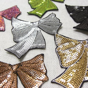 May include: Assortment of six sequined bow appliques in various colours, including silver, gold, pink, green, and brown. The bows are made of sequins and have a shiny, sparkly finish.
