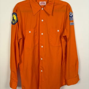 May include: Bright orange long-sleeve shirt with two chest pockets and white buttons. Patches on the sleeves read "Medical Technician" and "Idaho Rescue Support". The shirt has a collar and is hanging on a black hanger.