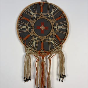 May include: A round, handmade dreamcatcher with a dark gray center and a wooden hoop. The design features orange, beige, and white woven patterns, with hanging tassels and black beads. This decorative piece is designed for wall decor.