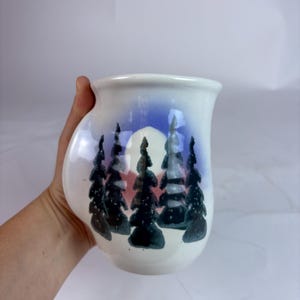 May include: White ceramic mug with a landscape design featuring dark green trees, a red and white sun, and a blue sky. The mug has a curved handle and a wide opening. The mug is held by a hand.