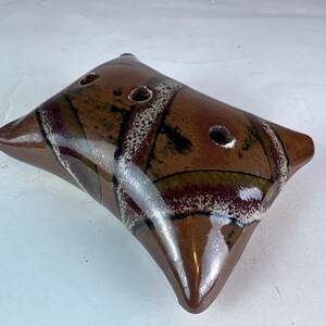 May include: A rectangular ceramic soap dish with a glossy brown glaze featuring abstract bands of dark red, olive green, and speckled white. The dish has a slightly concave surface with three circular drainage holes arranged in a row.