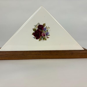May include: A triangular napkin holder with a white surface, adorned with a floral bouquet of burgundy roses, purple, and blue flowers. The base is made of dark wood. A decorative item for the dining table.