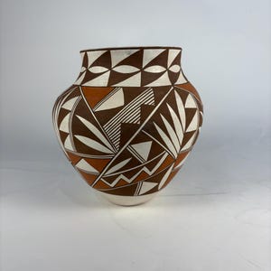 May include: A handcrafted pottery vase with a wide body and a narrow neck. The vase features a geometric design in brown, white, and orange. The pattern includes triangles, lines, and leaf-like shapes. The vase is likely used for decorative purposes.