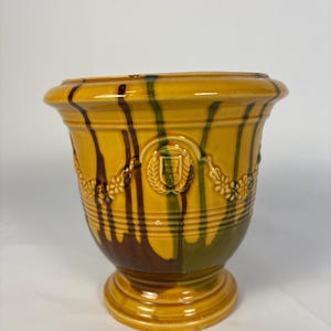 May include: A yellow ceramic planter with a pedestal base. The planter features a decorative design with brown and green vertical stripes, a floral garland, and a crest. The planter is suitable for indoor or outdoor use.